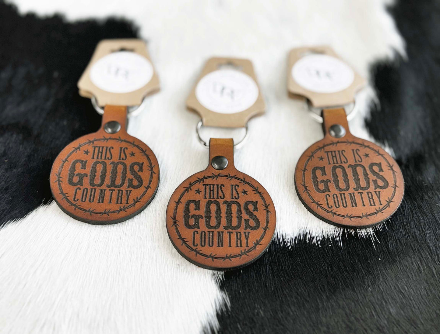 Three leather keychains with the text 'GOD'S COUNTRY' stamped on them, displayed on a furry white background.