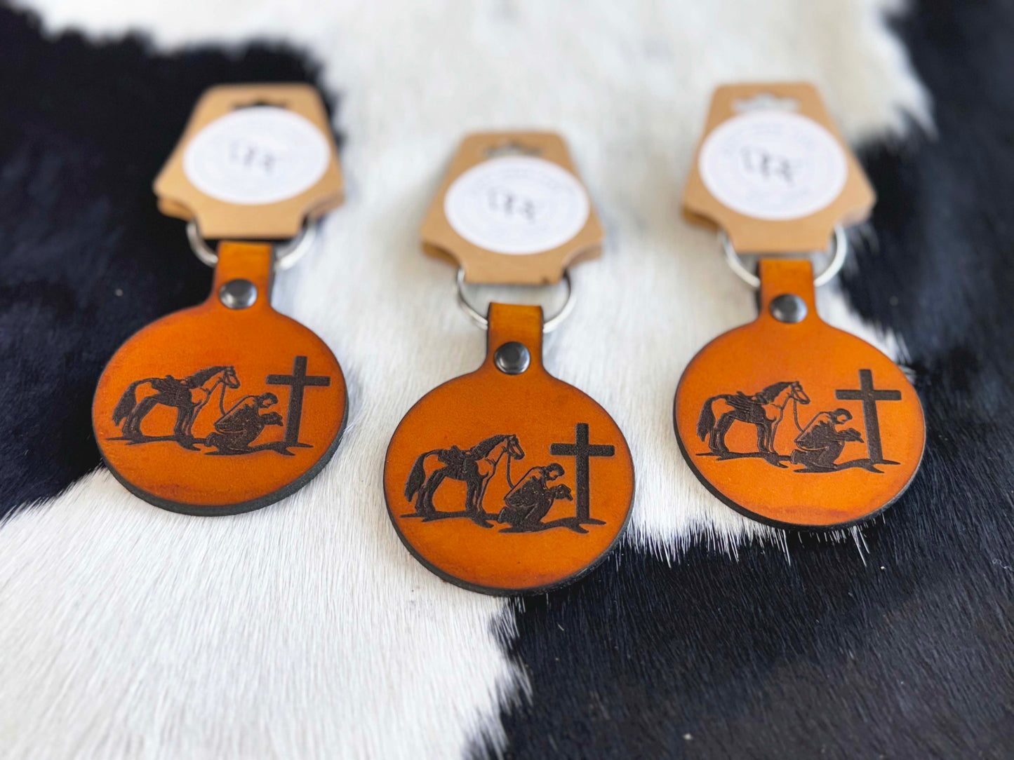 Three leather keychains with a cowboy and a cross design, displayed on a furry surface.