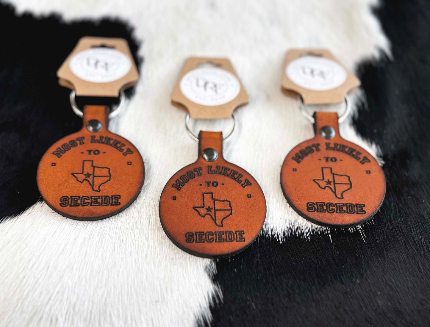 Three brown leather keychains with text and design related to the state of Texas, displayed on a furry surface.