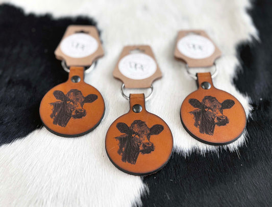 Three leather keychains with a cow head design on a brown background, displayed on a furry surface.