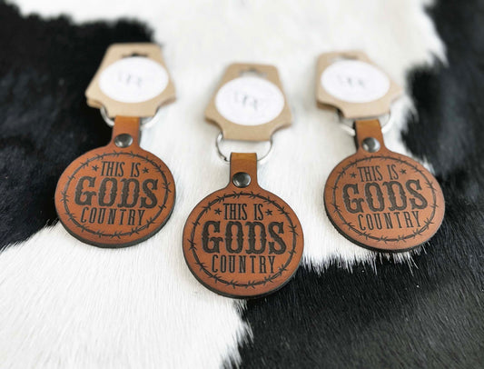 Three leather keychains with the text 'GOD'S COUNTRY' stamped on them, displayed on a furry white background.
