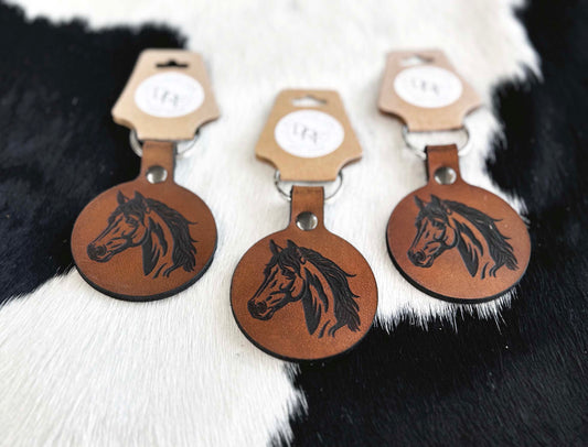 Three leather keychains with horse silhouette design, laid out on a black and white furry surface.