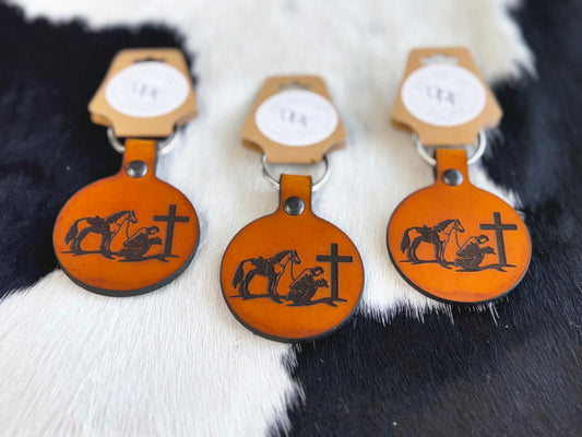 Three leather keychains with a cowboy and a cross design, displayed on a furry surface.