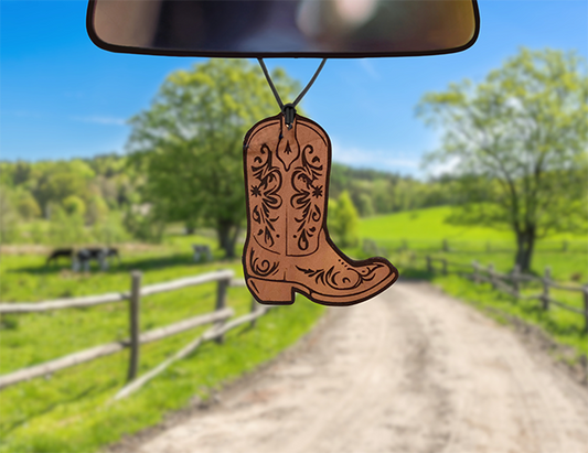 A leather car freshener in the shape of a boot hanging from a car's rearview mirror with a rural road and landscape in the background.