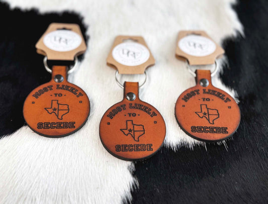 Three brown leather keychains with text and design related to the state of Texas, displayed on a furry surface.