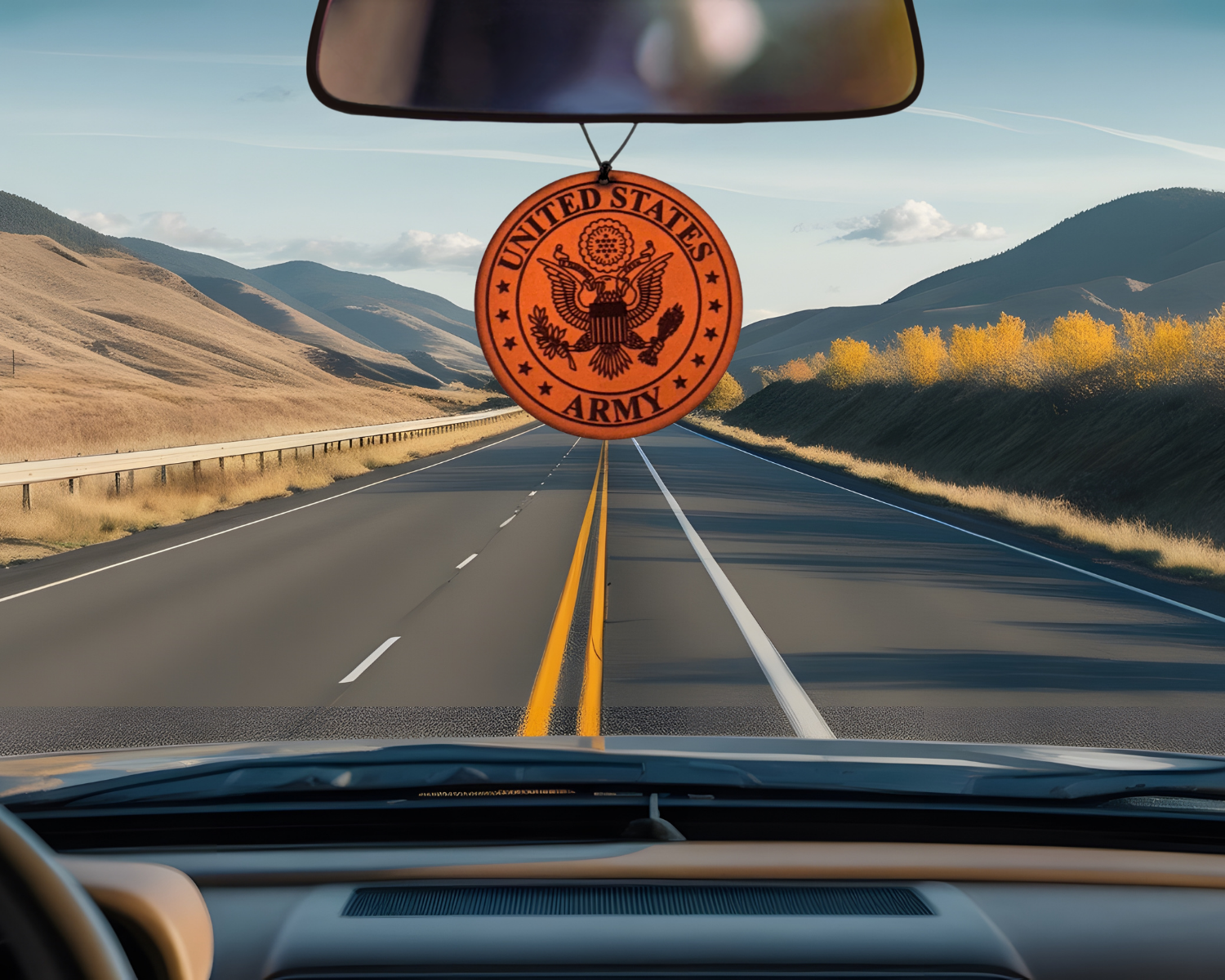 United States Army emblem hanging from a car's rearview mirror with a scenic road in the background.